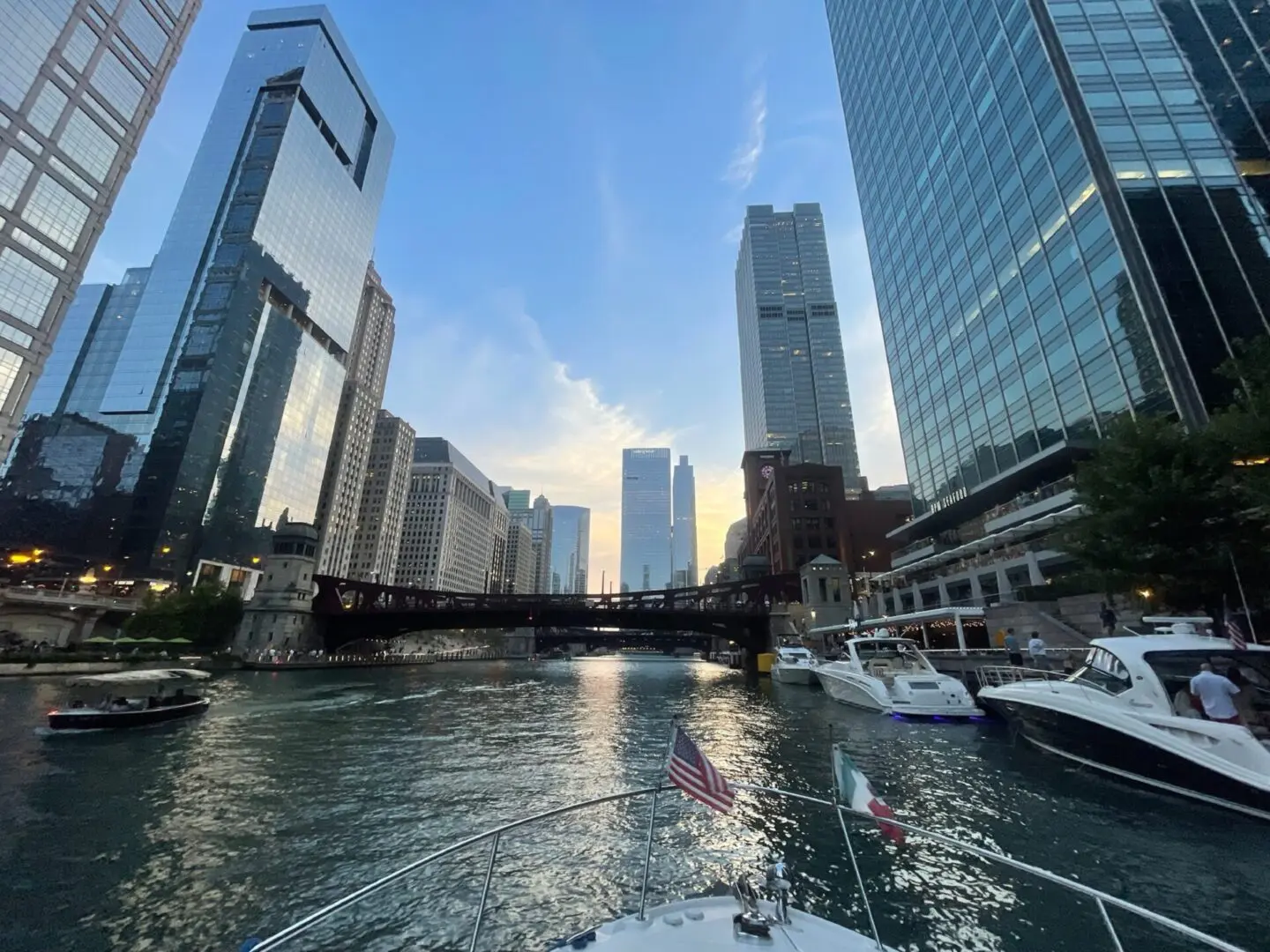 71288971720__2DCD434B-AE6B-4D19-ACA3-293C9AE79904.fullsizerender_page-0001 (1) View from a boat on a river with modern skyscrapers on both sides and a bridge in the distance under a blue sky at sunset.