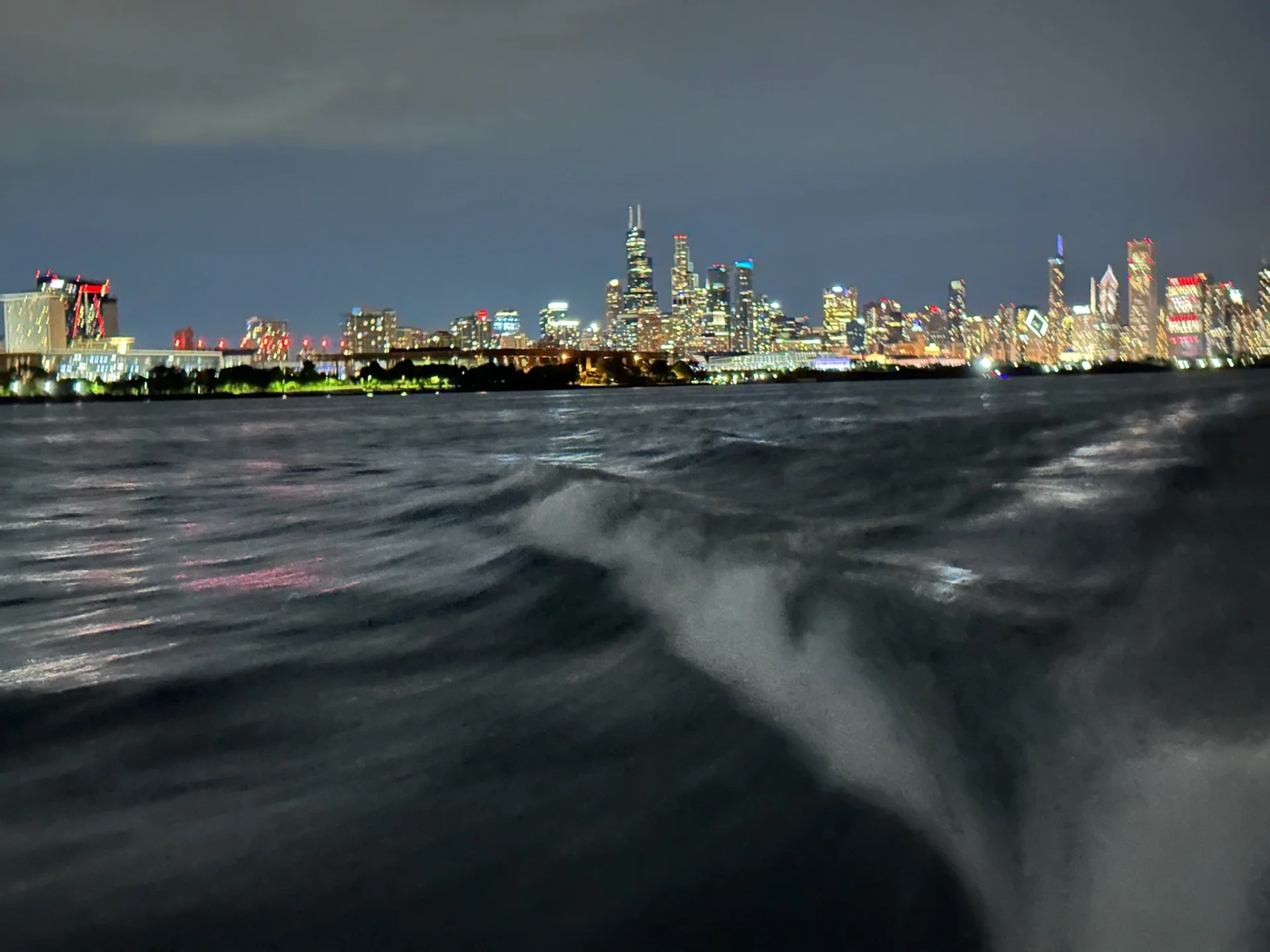 Nighttime view of a city skyline with bright lights from buildings reflecting on the water, photographed from a moving boat.